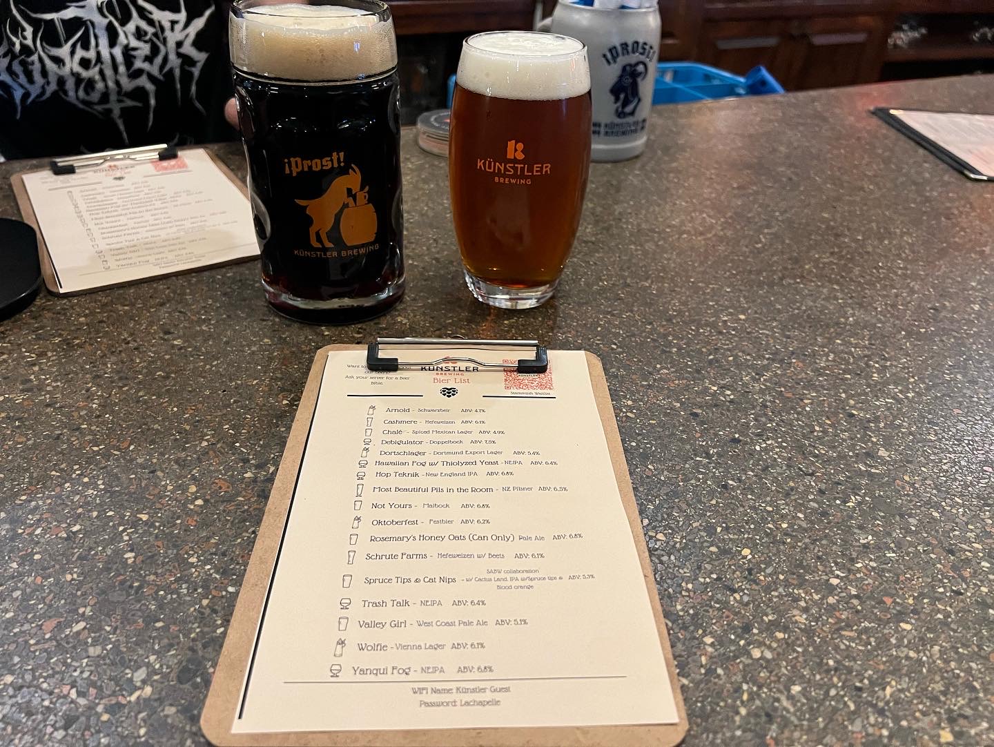 Two beers on a bar counter at Künstler Brewing—one dark beer in a branded pint glass and one amber beer in a stemmed glass—alongside a beer menu on a clipboard.