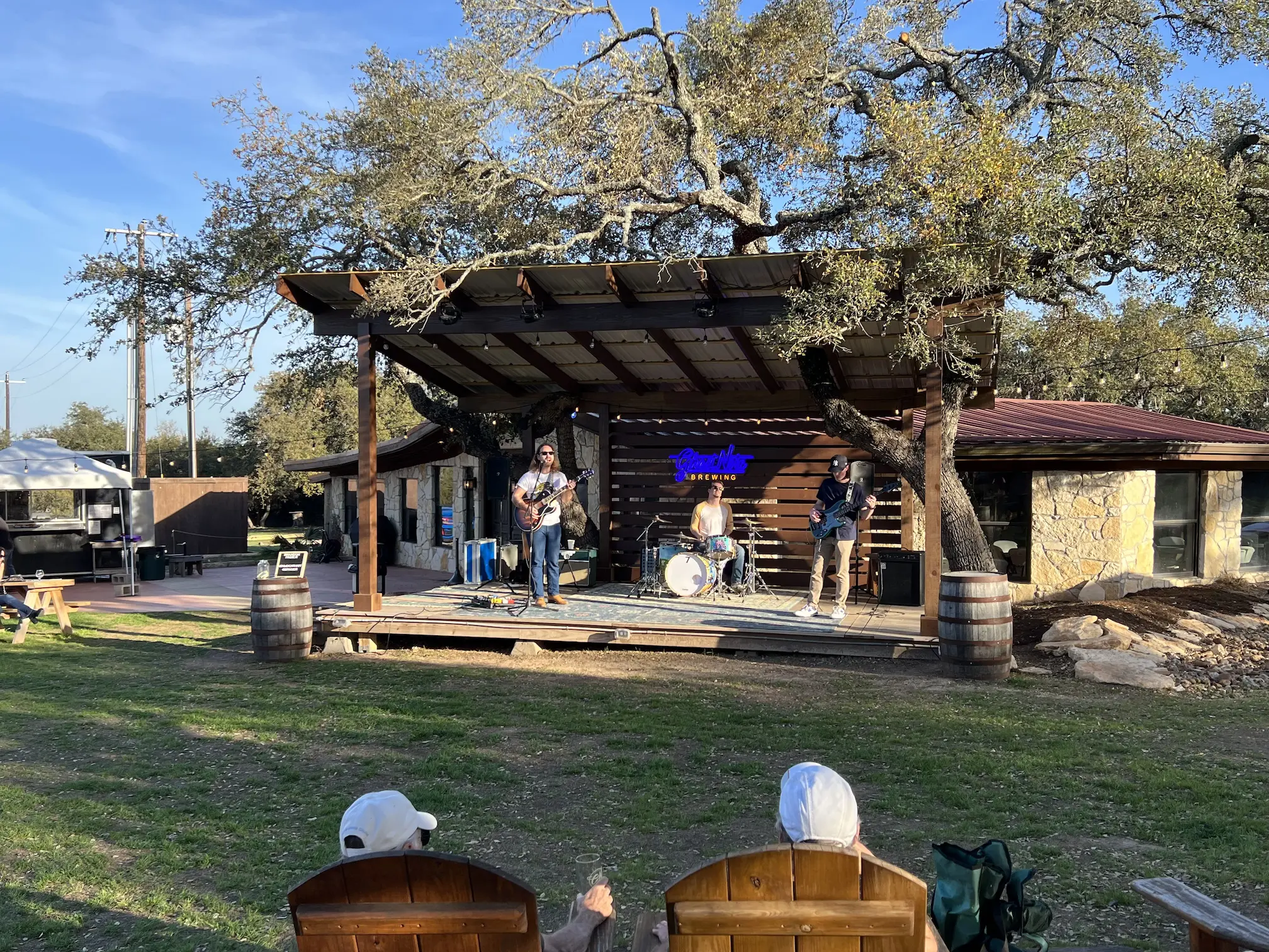 Live band performing on an outdoor wooden stage surrounded by oak trees at Ghost Note Brewery, with audience members seated in wooden chairs on a grassy lawn.