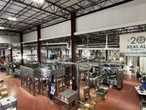 Interior view of Real Ale Brewing Co.'s production facility, showcasing large stainless steel brewing equipment, conveyor systems, and industrial machinery under a high metal ceiling.