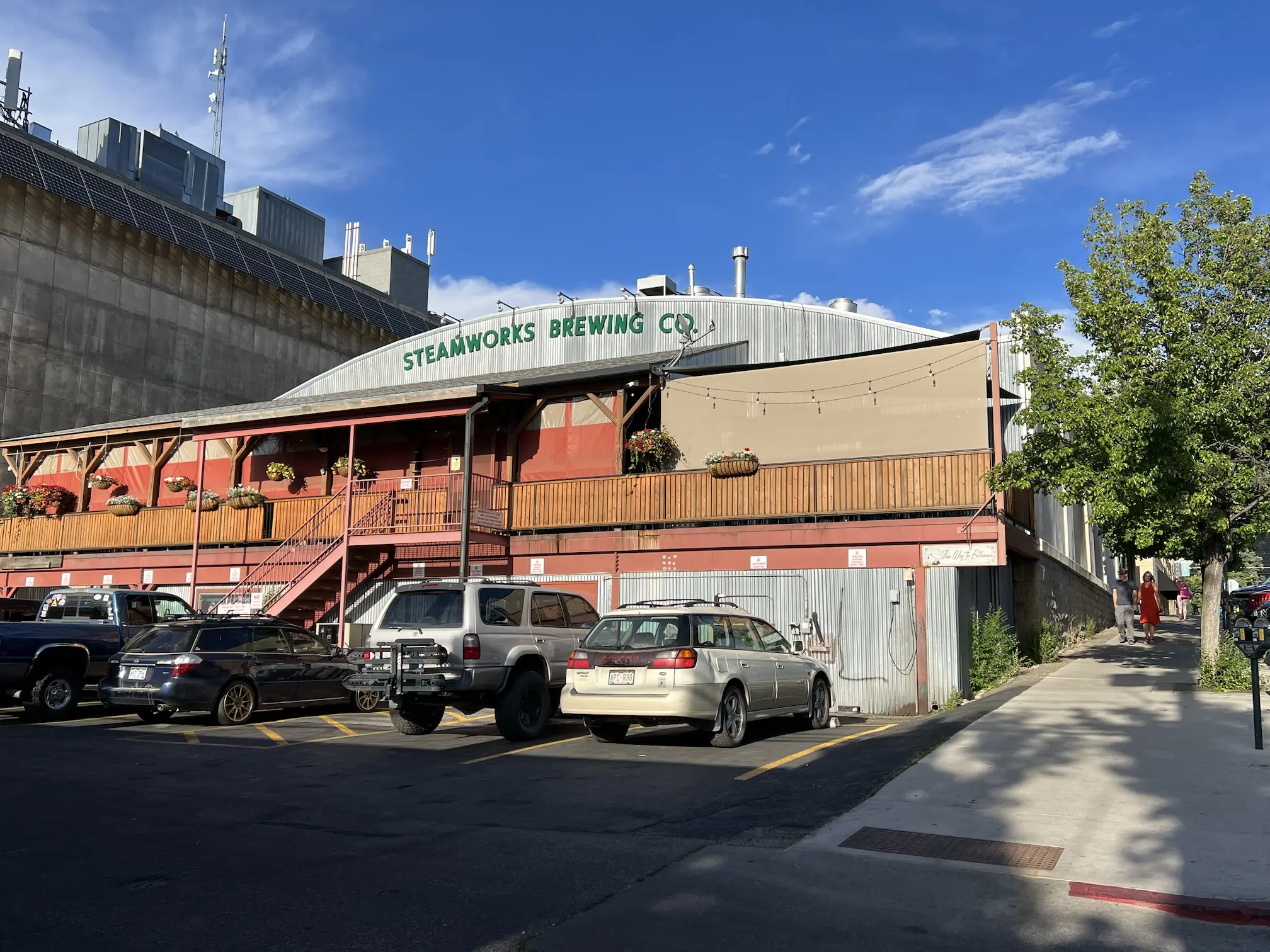 Exterior view of Steamworks Brewing Co. with a red and grey industrial-style building, outdoor patio with string lights, and parked cars in front under a clear blue sky.