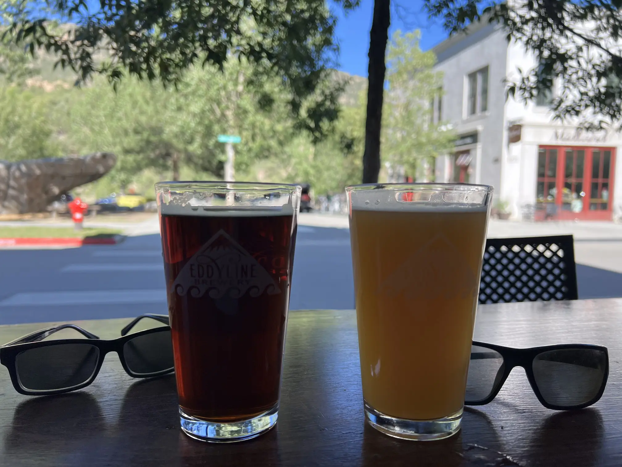 Two pints of beer—a dark amber and a hazy pale ale—sitting on a wooden table outside Eddyline Brewery at SouthMain in Buena Vista, Colorado, with sunglasses and a tree-lined street in the background.