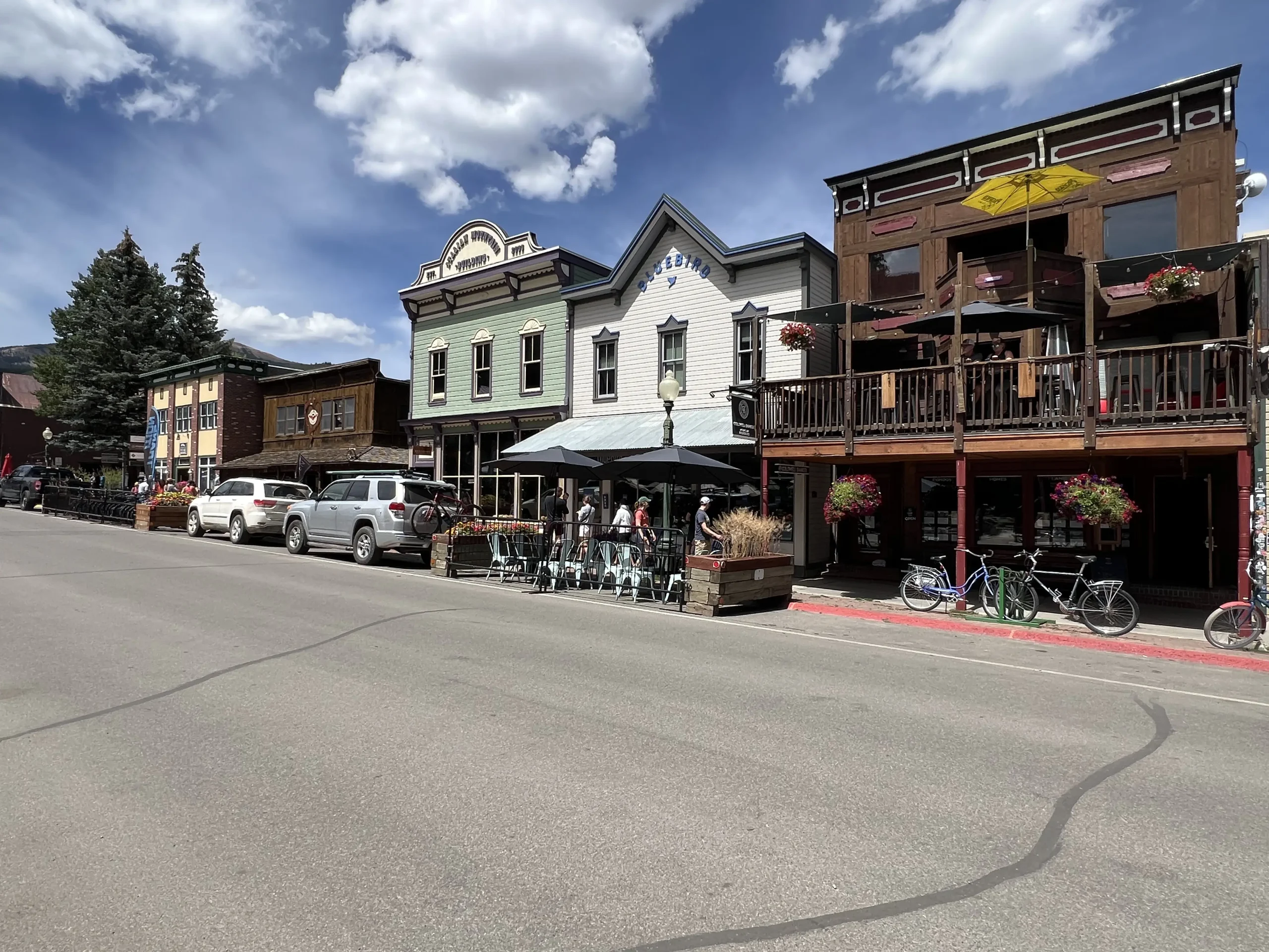 The Eldo Brewery and Taproom in Crested Butte, Colorado, showcasing its rustic wooden exterior, outdoor patio seating with umbrellas, bicycles parked nearby, and vibrant hanging flower baskets under a clear blue sky.