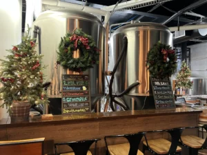 Holiday-decorated stainless steel brewing tanks behind a small bar at New Braunfels Brewery, complete with festive wreaths, a lit Christmas tree, and a chalkboard listing various craft beers.