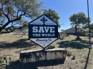 Sign for Save the World Brewing Co. in Marble Falls, Texas, with a scenic background of trees and the brewery building visible in the distance.