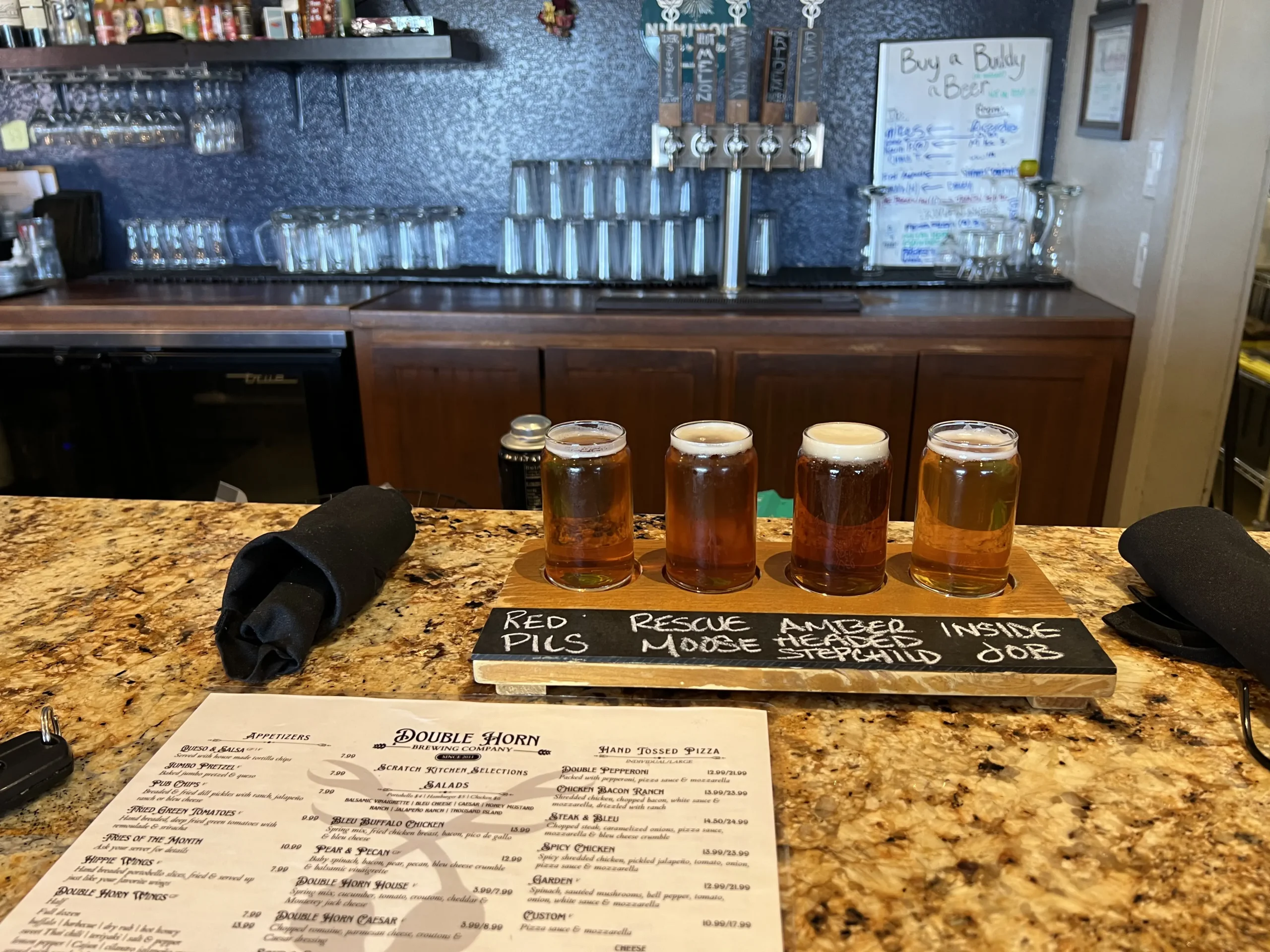 Photo of a flight of four craft beers on a wooden paddle labeled with beer names: Red Pils, Rescue Amber, Moose Inside, and Stepchild Job, placed on a granite bar at Double Horn Brewing Company. The background shows beer taps, glassware, and a menu on the counter.