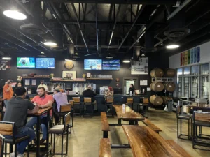 Interior view of Bold Republic Brewing showing patrons seated at wooden tables, a bar lined with multiple beer taps, TV screens overhead, and barrels stacked on the right.