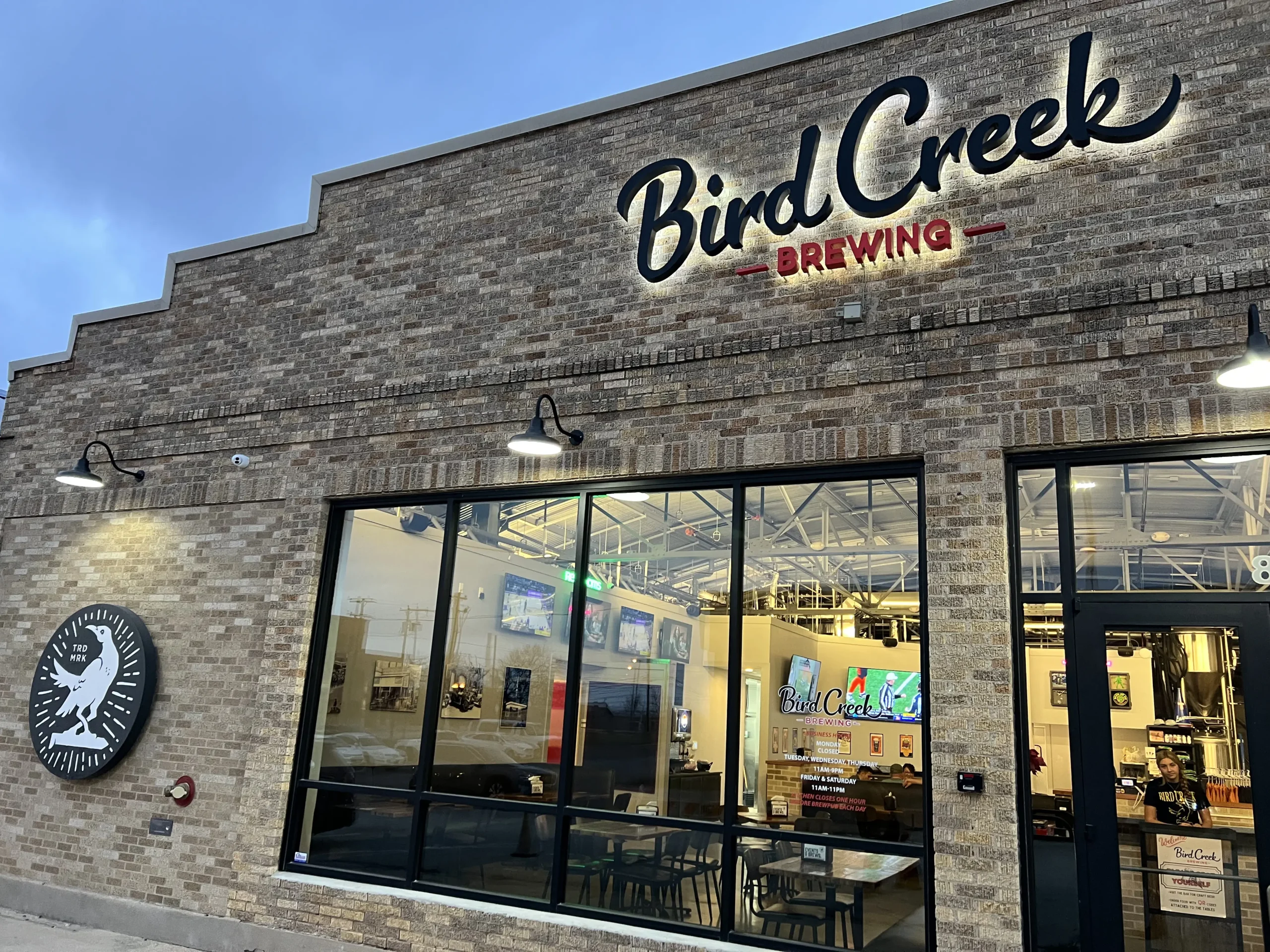 Front exterior view of the Bird Creek Brewing building with a brick facade, illuminated signage reading ‘Bird Creek Brewing,’ and large windows showing the taproom interior.